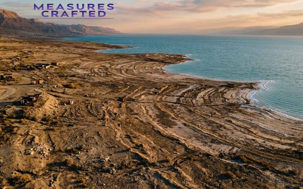 Aerial view of circular patterns in dry earth bordering the turquoise Dead Sea. Arid cliffs and sunset sky in the background.