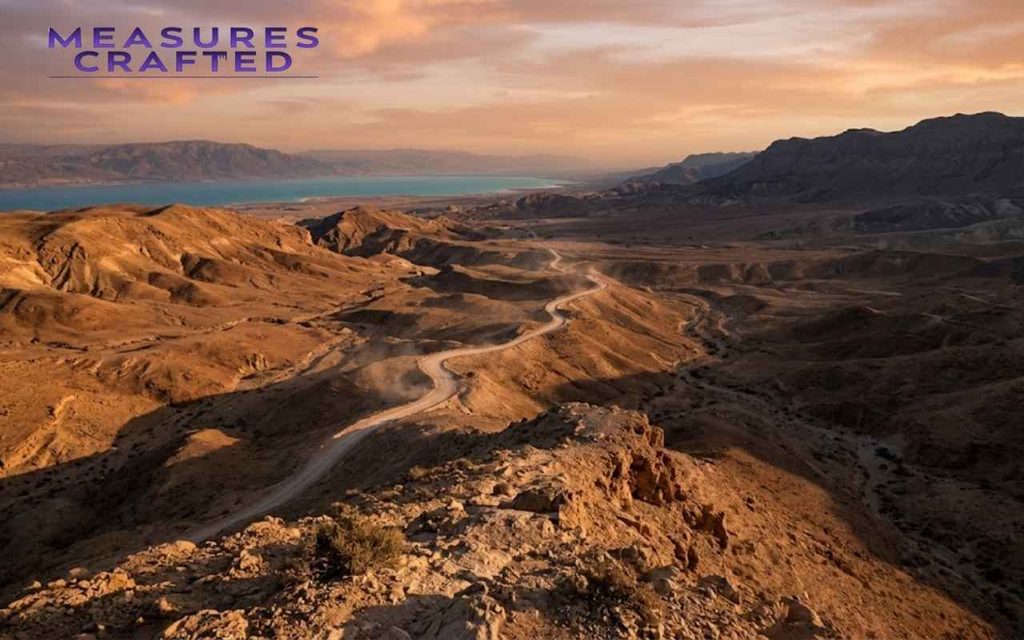 Winding dirt road through a vast, arid desert landscape. The turquoise Dead Sea and mountain range at sunset.