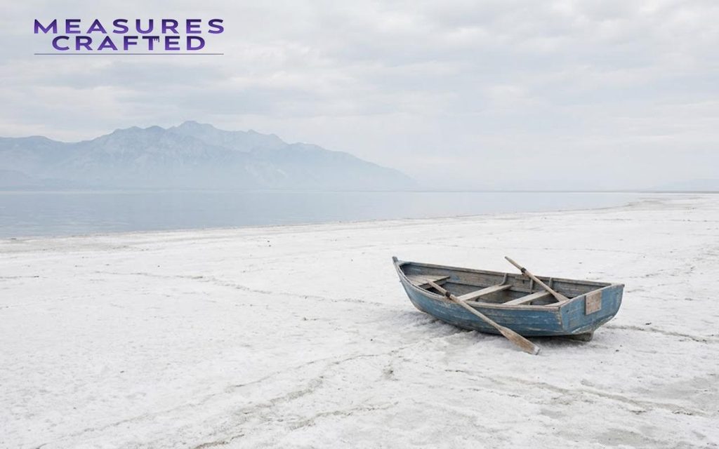 An blue rowboat sits isolated on a wide, cracked white salt flat. Beyond the flats are calm lake and hazy mountains.