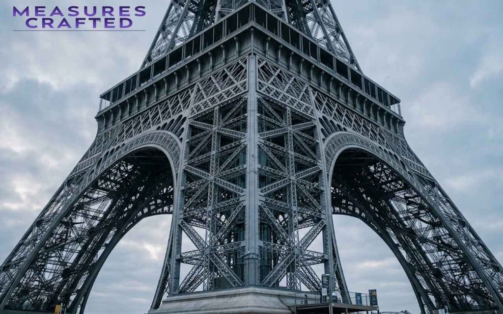Intricate iron latticework and arched base of the Eiffel Tower viewed from below against an overcast morning sky.