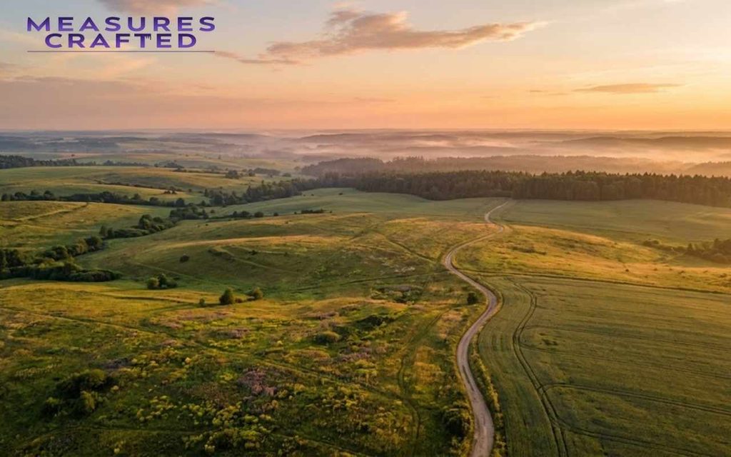 Aerial view of rolling 100 acres green meadows and a winding dirt path glowing under golden hour sunlight.