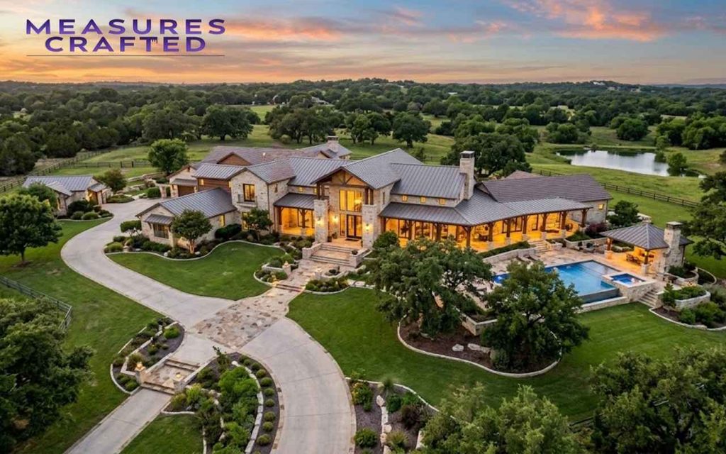 Aerial view of a luxury stone ranch estate with a pool, driveway, and lush grounds at sunset.