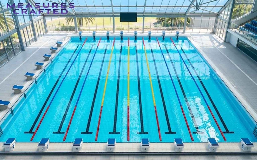 An indoor Olympic-sized swimming pool with turquoise water, red and white lane lines, and starting blocks under a glass roof.