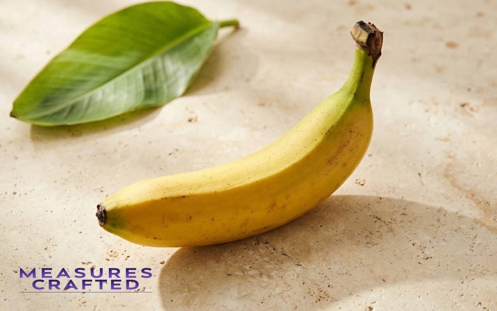 A single banana with natural speckles on warm cream stone next to a green leaf.